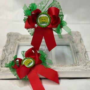 May include: Two red hair bows with green accents and a Grinch-themed centre. Each bow features a red ribbon, green lace, and a circular emblem with the word "Grinch". A white pearl is attached to one bow.