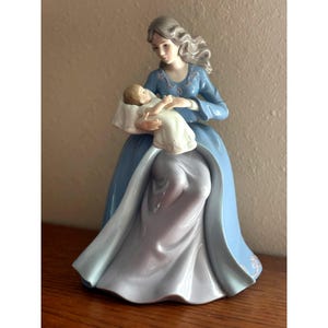 May include: A porcelain figurine of a woman in a long blue dress holding a baby wrapped in white cloth. The woman has gray hair and is looking down at the baby. The figurine is on a wooden surface.
