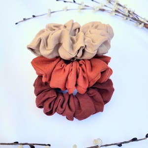 May include: Three fabric scrunchies stacked on top of each other. The top scrunchie is beige, the middle scrunchie is rust orange, and the bottom scrunchie is a darker shade of rust orange.