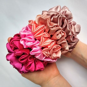 May include: A set of five satin scrunchies in shades of pink, peach, and red. The scrunchies are arranged in a bouquet shape and are held in a hand.