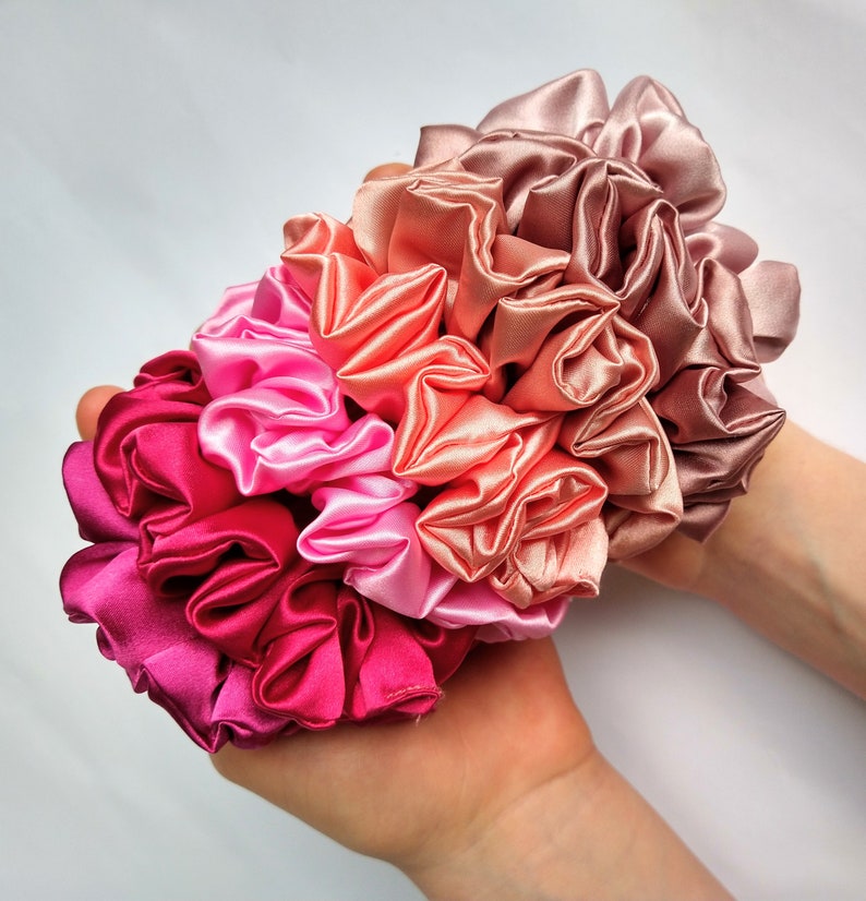 May include: A set of five satin scrunchies in shades of pink, peach, and red. The scrunchies are arranged in a bouquet shape and are held in a hand.