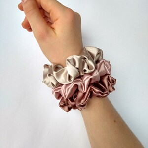 May include: Three stacked scrunchies in shades of pink and beige. The scrunchies are made of soft, silky fabric and are perfect for holding hair in place.