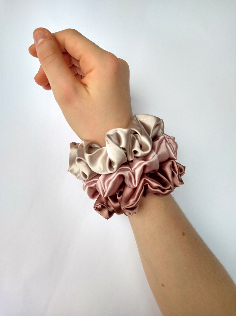 May include: Three stacked scrunchies in shades of pink and beige. The scrunchies are made of soft, silky fabric and are perfect for holding hair in place.