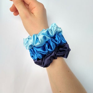 May include: A set of three scrunchies in shades of blue, from light blue to dark blue. The scrunchies are made of satin fabric and are stacked on top of each other.