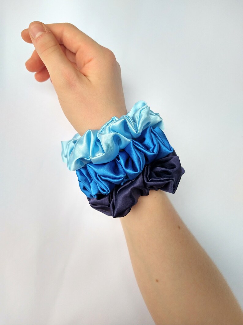 May include: A set of three scrunchies in shades of blue, from light blue to dark blue. The scrunchies are made of satin fabric and are stacked on top of each other.