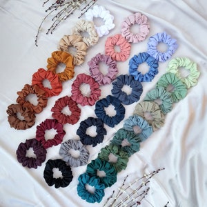May include: A collection of 20 colourful scrunchies made of fabric. The scrunchies are arranged in rows, with the colours transitioning from light to dark. The scrunchies are all made of the same type of fabric, but they are in different colours, including white, light pink, lavender, light green, dark green, blue, red, orange, brown, and black.