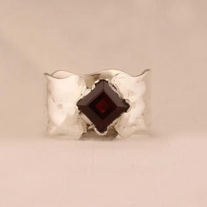 May include: A silver ring with a textured band and a square-shaped, dark red gemstone.