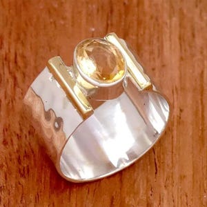 Könnte beinhalten: Ein breiter Silberring mit gehämmertem Muster, besetzt mit einem großen, ovalen, goldgelben Edelstein. Der Ring hat goldfarbene Akzente auf beiden Seiten des Edelsteins, was einen auffälligen Kontrast erzeugt.