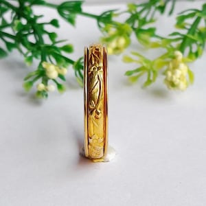 Pode incluir: Um anel de ouro com um padrão floral gravado. O anel é de uma cor dourada brilhante e brilhante e apresenta entalhes detalhados de folhas e flores ao redor da banda. O anel é colocado sobre um fundo branco com folhagem verde.