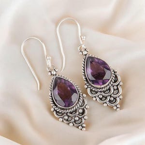 Könnte beinhalten: Ein Paar Ohrhänger aus Sterlingsilber mit facettierten, birnenförmigen violetten Amethysten. Die Silbereinfassungen verfügen über aufwendige Kordelränder und dekorative, geschwungene Metallarbeiten mit kleinen Silberkügelchen an der Unterseite.