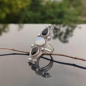 Könnte beinhalten: Ein silberner Ring mit drei tropfenförmigen schwarzen Edelsteinen und einem zentralen runden, irisierenden Mondstein. Der Ring hat ein verziertes, detailliertes Design mit geteiltem Schaft. Der Ring wird auf einer reflektierenden Oberfläche präsentiert.