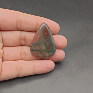 Könnte beinhalten: Ein tropfenförmiger Cabochon aus grünem, rotem und braunem Jaspis. Der Stein hat eine marmorierte Optik mit Farbwirbeln.