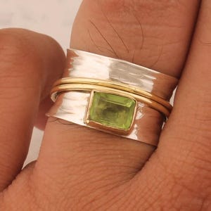 May include: A silver and gold ring with a green gemstone. The ring is a wide band with a hammered texture. The gemstone is a square cut and is set in a gold bezel.