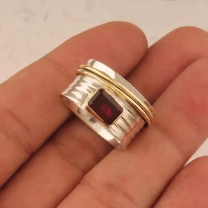 May include: A silver ring with a rectangular, deep red gemstone. The ring has a textured band and a thin gold band encircling the top. The ring is a piece of jewelry.