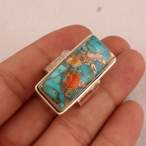 Könnte beinhalten: Ein silberner Ring mit einem rechteckigen Türkis-Stein, der in der Mitte eingesetzt ist. Der Stein hat eine Mischung aus blauen, grünen, orangen und weißen Farben.