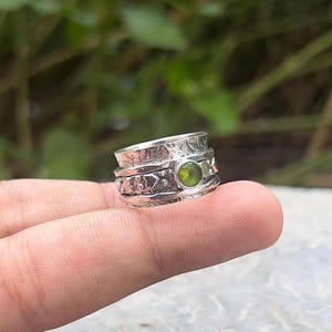 Könnte beinhalten: Ein silberner Spinner-Ring mit einem grünen Edelstein. Der Ring hat ein strukturiertes Design mit floralen und Herzmustern. Der Edelstein ist rund und in der Mitte des Rings gefasst. Der Ring wird von einem Finger gehalten.
