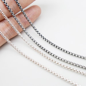 May include: Four box chain necklaces in silver, gray, and black. The chains are displayed against a white background. The chains are made of small, square links.