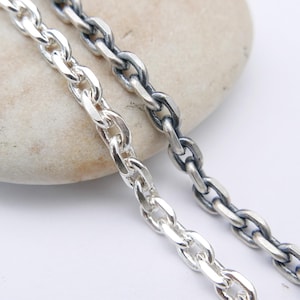 May include: Two silver chain necklaces with oval links, one polished and bright, the other oxidized to a darker gray. The chains are displayed on a light-colored, smooth stone against a white background. Jewelry.