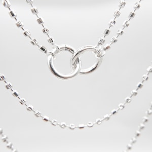 Może przedstawiać: Zbliżenie trzech srebrnych naszyjników. Naszyjniki mają delikatne łańcuszki kulkowe i dwa zazębiające się pierścienie. Łańcuszki zawieszone są na miękkim, białym tle, podkreślając połysk i design biżuterii.