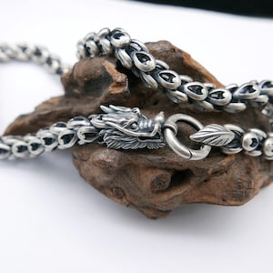 May include: A silver-colored dragon-themed necklace with a detailed dragon head clasp. The chain is made of interconnected oval links. The necklace rests on a piece of dark, textured wood against a white background. The jewelry is likely made of sterling silver.