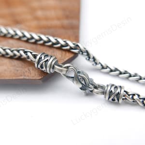 Könnte beinhalten: Eine silberfarbene Halskette mit geflochtenem Design. Die Halskette hat dekorative Verschlüsse mit komplizierten Mustern und einen detaillierten Verschluss. Die Kette wird auf einer Holzoberfläche präsentiert.