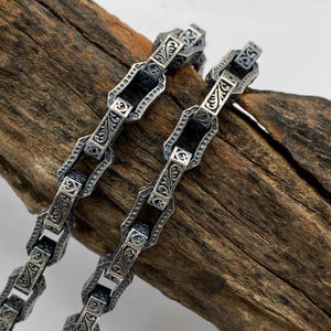 May include: Close-up of two silver chains with ornate, engraved links. The chains have a detailed, vintage design with repeating patterns. The chains are displayed on a piece of weathered wood, highlighting the contrast between the metal and the natural texture.