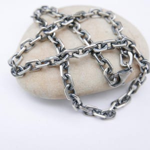 Puede incluir: Un collar de cadena plateada con un diseño de eslabones gruesos, apoyado sobre una piedra lisa de color claro. El collar se muestra sobre un fondo blanco, resaltando su brillo metálico y sus intrincados detalles. Los eslabones de la cadena están claramente definidos.