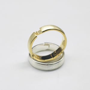 Könnte beinhalten: Zwei Eheringe, einer aus Gold und einer aus Silber, sind gestapelt. Der goldene Ring hat ein einzigartiges Design mit einer kleinen Lücke, während der silberne Ring ein einfaches Band ist. Die Ringe sind vor einem schlichten weißen Hintergrund platziert.