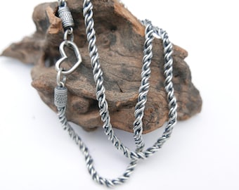 May include: A silver-toned necklace with a heart-shaped pendant and a twisted rope chain. The necklace is displayed on a piece of weathered wood against a white background. The heart pendant adds a touch of romance.