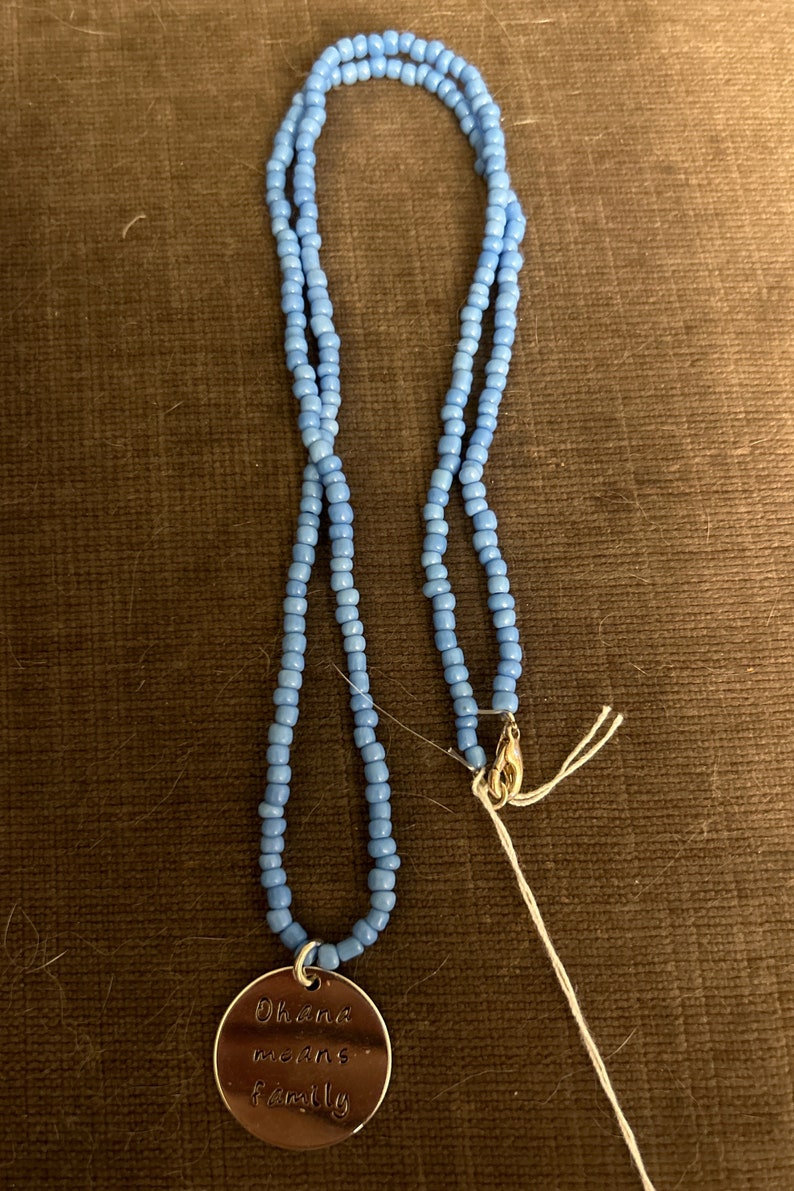Puede incluir: Un collar de cuentas azul claro con un dije de plata que dice "Ohana means family".
