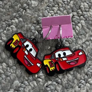 May include: A pair of red Lightning McQueen earrings. Each earring features a smiling cartoon car with black outlines, white eyes, and yellow accents. The earrings are attached to silver-colored ear wires and a pink earring display card.