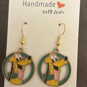 May include: A pair of gold-colored dangle earrings featuring a yellow cartoon dog with a green border. The dog has a pink tongue and black accents. The earrings are on a white card with the text "Handmade with love".