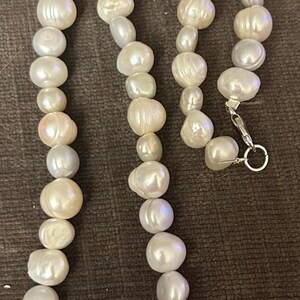 Op de afbeelding: Een ketting bestaande uit onregelmatig gevormde, gebroken witte parels. De parels variëren in grootte en hebben een glanzende glans. Een zilverkleurige sluiting zet de ketting vast. De ketting wordt gepresenteerd op een bruin oppervlak.
