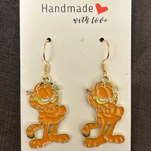 Könnte beinhalten: Ein Paar goldene Ohrhänger mit einem Cartoon-Bild von Garfield der Katze. Die Ohrringe sind handgefertigt mit Liebe.