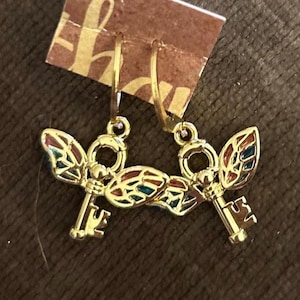May include: Gold-toned earrings featuring a key design with colorful enamel wings. The keys have intricate details and are suspended from hoop closures. The wings are filled with red, blue, and green enamel. The earrings are displayed against a brown background.