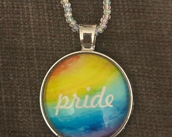 Puede incluir: Un collar de plata con un colgante redondo que presenta un diseño de arcoíris de acuarela y la palabra "pride" en letras blancas.