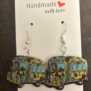 May include: A pair of school bus earrings with silver hooks. The earrings are yellow with a cartoon bus design, featuring a driver and musical instruments. The card behind the earrings reads "Handmade with love" with a red heart.