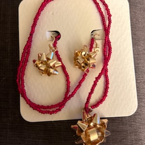 Peut inclure: Un collier de perles rouges avec un pendentif en forme de nœud doré et des boucles d'oreilles assorties. Le collier et les boucles d'oreilles sont présentés sur une carte blanche.