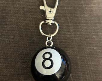 May include: A black and silver eight ball keychain with a silver clasp. The number eight is prominently displayed in white on a silver circle. The keychain is attached to a silver metal clasp.