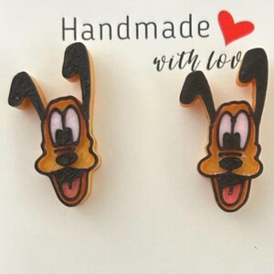 May include: Handmade Pluto dog earrings. These earrings feature a cartoon Pluto dog design with orange, black, and white colors. The earrings are shaped like Pluto's head and have a tongue sticking out. The text "Handmade with love" is above the earrings.