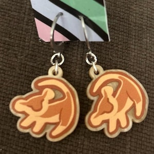 May include: A pair of dangle earrings featuring a Simba design. The earrings have silver-colored hoops and are made of a light-colored material with an orange and brown Simba design. The earrings are hanging against a brown background.