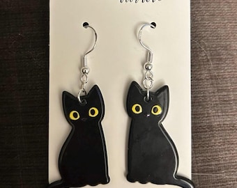 May include: A pair of black cat earrings with yellow eyes and silver-colored hooks. The earrings are on a white card with the text "Handmade with love" and a red heart.