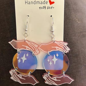 May include: A pair of handmade earrings with a celestial theme. Each earring features a silver hook, a hand-shaped charm, and a circular pendant with a blue square and star design. The text "Handmade with love" is visible.