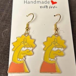 May include: A pair of gold-colored dangle earrings featuring a yellow cartoon character with an orange dress. The earrings are made with a gold-colored hook and a yellow charm. The earrings are on a white card with the text "Handmade with love".