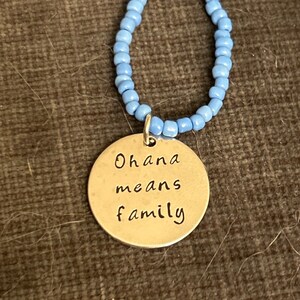 Puede incluir: Un collar de plata con un colgante redondo que dice "Ohana means family". El collar est&aacute; hecho de cuentas de color azul claro.