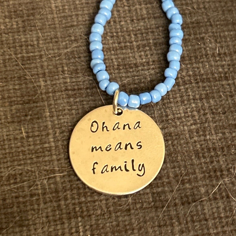 Puede incluir: Un collar de plata con un colgante redondo que dice "Ohana means family". El collar est&aacute; hecho de cuentas de color azul claro.