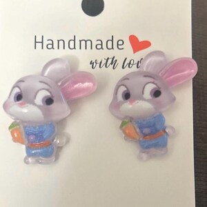 May include: A pair of stud earrings featuring a cartoon rabbit character with light purple fur, pink inner ears, and a blue uniform, holding an orange carrot. The earrings are attached to a white display card with the text Handmade with love.