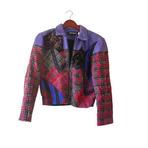 May include: A vintage jacket with a unique patchwork design. The jacket features a purple collar and shoulders, with a mix of red plaid, black, and blue patterned fabrics. It has a full zipper and is hanging on a wooden hanger.