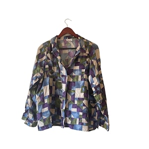 Pode incluir: Camisa abotoada com um padrão geométrico em tons de azul, roxo, verde e bege. A camisa está pendurada em um cabide de madeira sobre um fundo branco. Possui mangas compridas e gola.