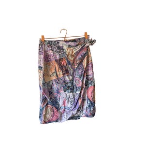 Peut inclure: Une jupe portefeuille avec un imprimé abstrait multicolore dans des tons de violet, rose, bleu et beige. La jupe présente une fermeture à nouer à la taille et est présentée sur un cintre en bois. Le motif inclut diverses formes géométriques.